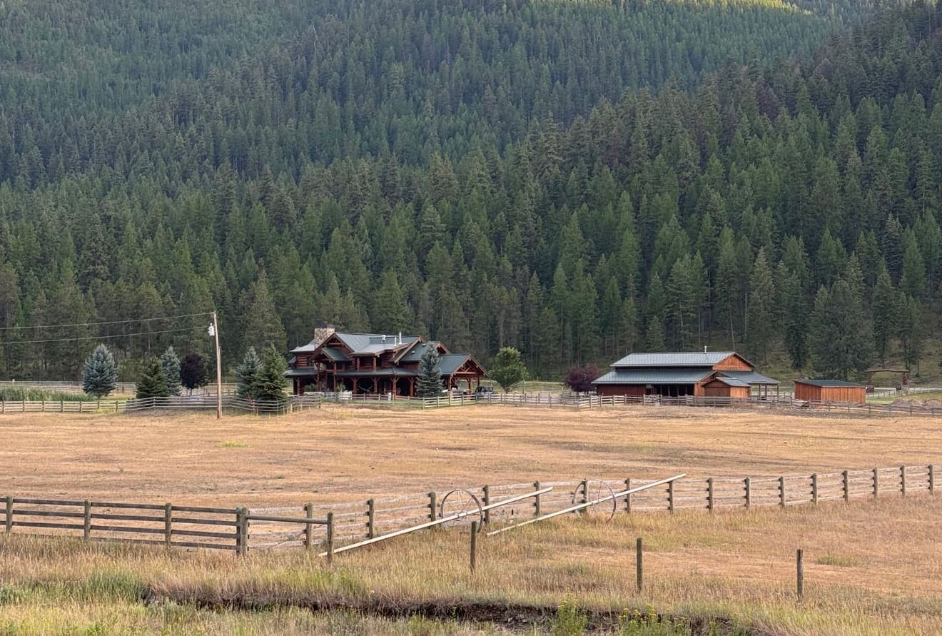 Ranch house and barn across dry late-summer pasture, with forested hillside behind